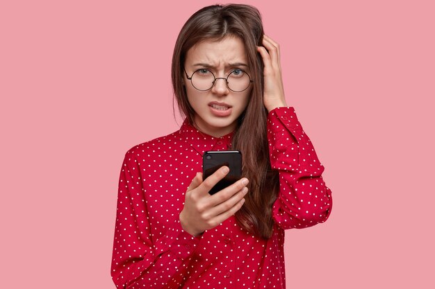 Foto de mujer de cabello oscuro disgustada mira con perplejidad, sostiene el teléfono móvil, reflexiona sobre el contenido del mensaje, vestida con ropa elegante