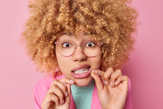 La foto de una mujer de cabello rizado limpia los dientes con hilo dental se encarga de la higiene bucal usa anteojos transparentes vestidos con ropa formal aislada sobre fondo rosa Concepto de salud dental