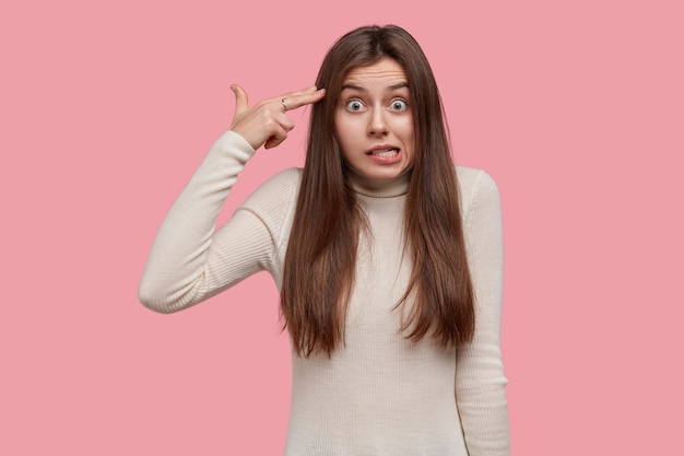 Foto de una mujer de Caucasain nerviosa y perpleja que hace un gesto de suicidio en la sien, dispara en la cabeza, tiene una expresión facial disgustada