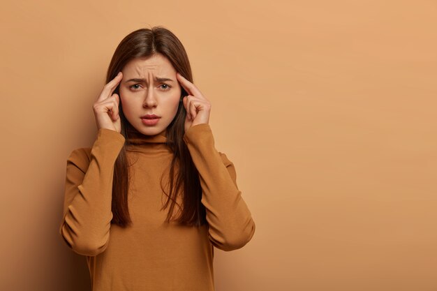 Foto de mujer caucásica intensa toca las sienes con los dedos índices, sonríe, sufre de dolor de cabeza o migraña, se siente enferma y molesta