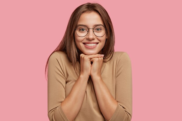 Foto de mujer caucásica morena de aspecto agradable tiene una sonrisa con dientes, sostiene la barbilla con ambas manos