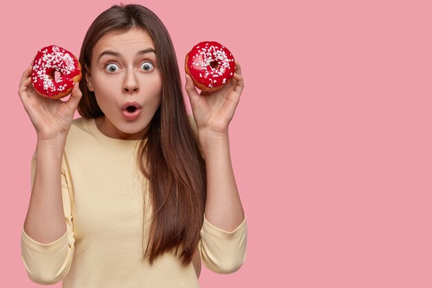 Foto de una mujer caucásica sorprendida contiene la respiración, lleva rosquillas dulces rojas