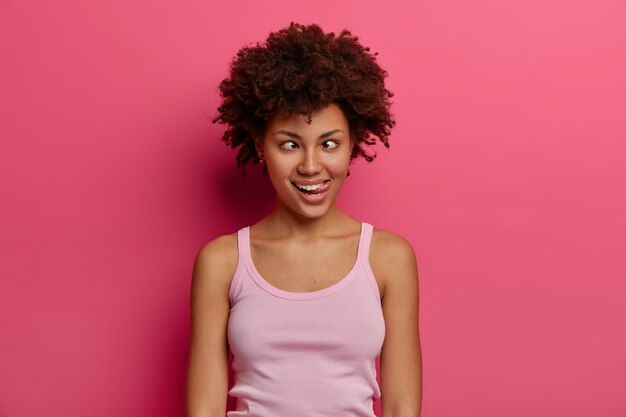 Foto de mujer cómica divertida que hace muecas, entrecierra los ojos y saca la lengua, hace el tonto, se divierte sola, está loca y tonta, vestida con ropa casual, posa contra la pared rosada. Estilo de vida fácil