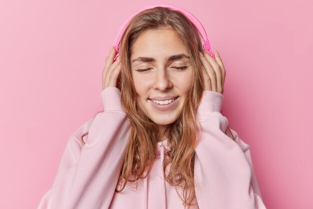 La foto de una mujer complacida de cabello largo mantiene los ojos cerrados, sonríe suavemente, usa capucha y disfruta de su pista favorita a través de auriculares inalámbricos aislados sobre fondo rosa. Hobby de personas y concepto de tiempo libre.