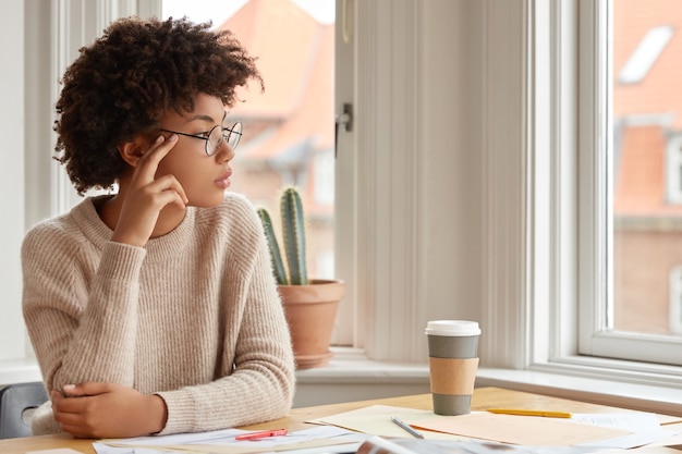 Foto de mujer contemplativa con peinado afro, usa gafas redondas, suéter cálido casual