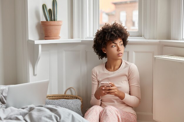 Foto de mujer contemplativa con peinado afro, viste ropa de dormir informal, comprueba la notificación en el teléfono inteligente, blogs en Internet