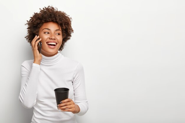 Foto de mujer despreocupada satisfecha con corte de pelo rizado, habla a través de un teléfono inteligente, mira positivamente a un lado, bebe café para llevar, está de buen humor durante una conversación animada. Personas y estilo de vida