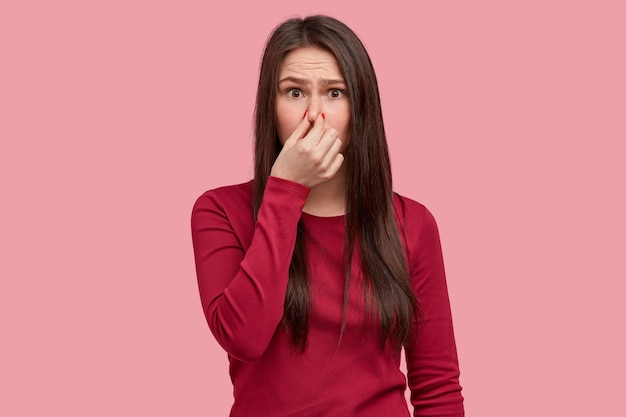 Foto de mujer disgustada que cierra la nariz con hedor, siente un olor terrible a basura, viste ropa roja