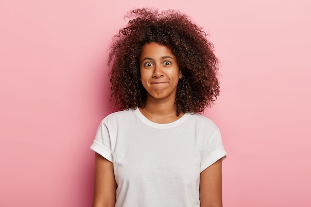 La foto de una mujer divertida tiene el pelo grueso y rizado, aprieta los labios, tiene una cara feliz, viste una camiseta blanca, aislada sobre una pared rosa. Buena chica afroamericana joven expresa felicidad.