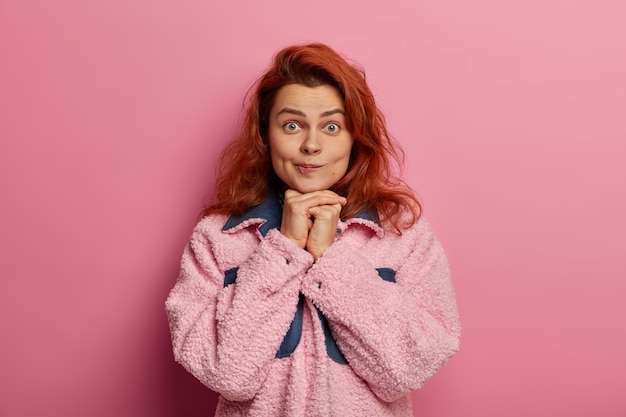 Foto de mujer encantadora con cabello pelirrojo natural, mantiene las manos debajo de la barbilla, frunce los labios, usa un abrigo rosado, mira con asombro, dice que no creo