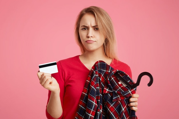 Foto de mujer encantadora con expresión de descontento va de compras en una boutique de moda, elige atuendo, sostiene una tarjeta de plástico