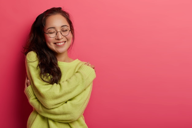 Foto de mujer encantadora que expresa amor propio y cuidado, disfruta vistiendo un nuevo suéter verde suave, abraza su propio cuerpo, recuerda un momento romántico con su novio