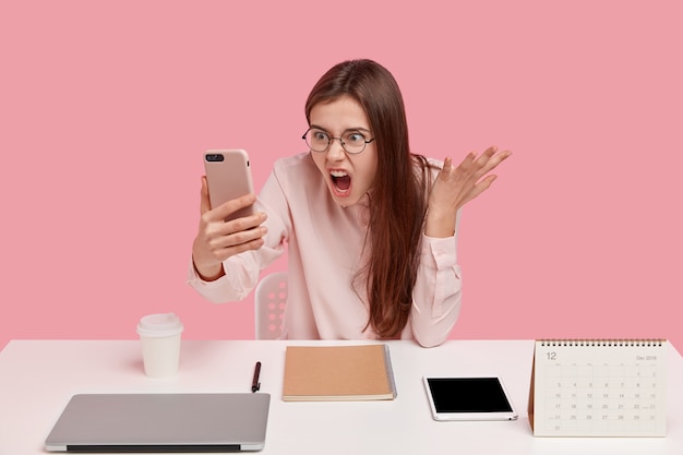 Foto de mujer enojada indignada sostiene un teléfono celular moderno, hace videollamadas, discute con un colega, posa en el lugar de trabajo