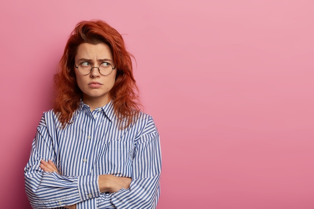 Foto de mujer enojada ofendida disgustada con mirada insultada y manos cruzadas, enfurruñado mientras mira hacia otro lado, usa gafas redondas y camisa a rayas azules