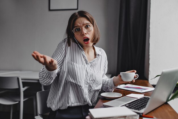 Foto de mujer enojada en vasos con taza de café en sus manos. Mujer de negocios mira fijamente a la cámara con asombro, hablando por teléfono en el lugar de trabajo.