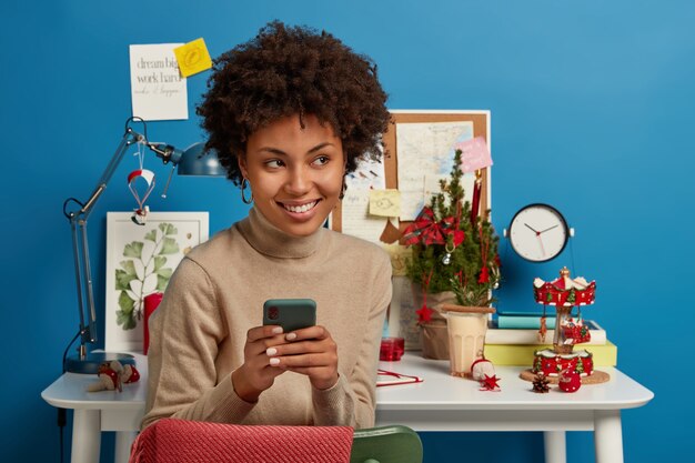 Foto de una mujer étnica autónoma que charla en línea en un teléfono inteligente, tiene un descanso después del trabajo, se sienta en la mesa cerca del escritorio, navega por Internet, se ve con una expresión feliz y soñadora a un lado, pared azul.