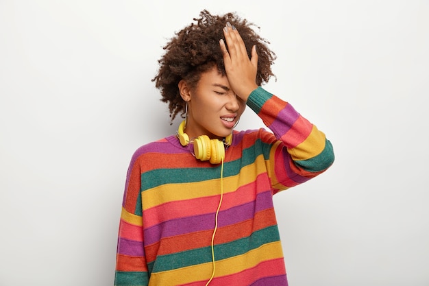 Foto de mujer étnica estresante golpea la frente con la palma de la mano, olvida información importante o se arrepiente de haber hecho algo mal, usa un jersey multicolor, auriculares