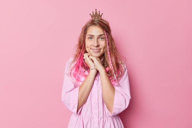 La foto de una mujer europea alegre y de buen aspecto finge ser princesa lleva corona en la cabeza y camisa sonríe tiernamente con hoyuelos en las mejillas mantiene la mano bajo la barbilla aislada sobre fondo rosa