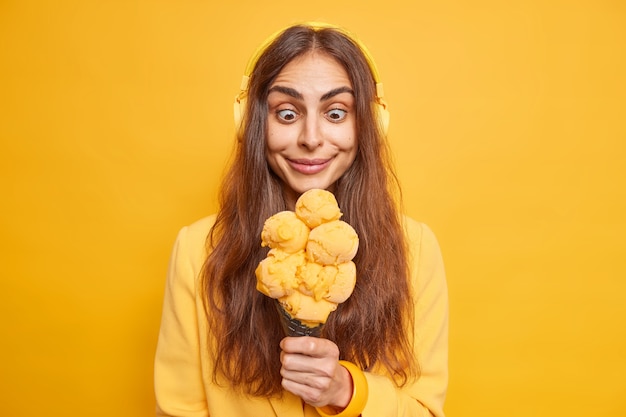 Foto de una mujer europea alegre y sorprendida con el pelo oscuro de lng que mira fijamente el apetitoso helado muere de hambre para comerlo escucha música a través de auriculares estéreo inalámbricos posa en el interior contra la pared amarilla