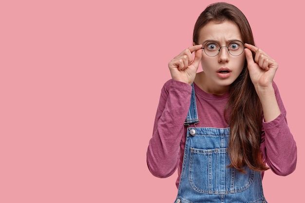 Foto de mujer europea joven asustada mantiene las manos en el marco de las gafas, tiene expresión de sorpresa, cabello largo