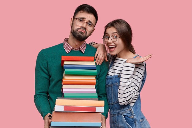 Foto de mujer europea positiva viste suéter a rayas y overoles, se inclina sobre el hombro del nerd masculino cansado con pila de libros gruesos