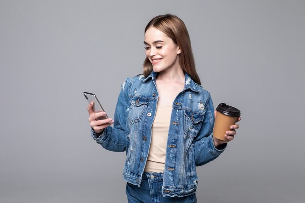 Foto de mujer exitosa de pie con smartphone y café para llevar en manos aisladas sobre pared gris