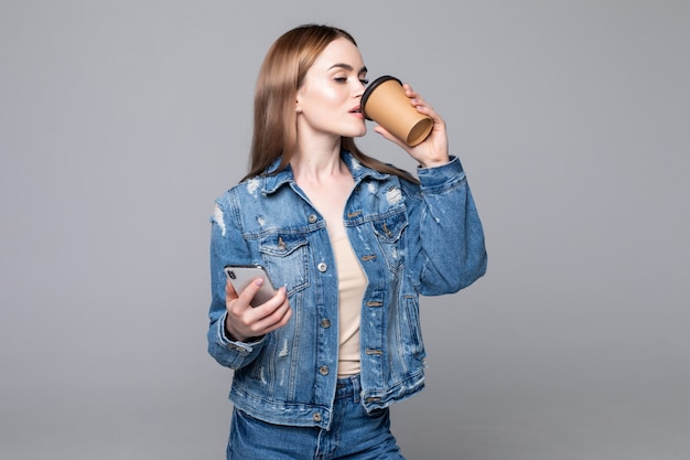 Foto de mujer exitosa de pie con smartphone y café para llevar en manos aisladas sobre pared gris