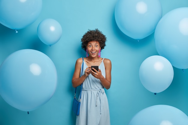 Foto de mujer feliz e impresionada en una fiesta corporativa, vestida de azul, sostiene un teléfono inteligente, se sorprende al recibir un mensaje inesperado de su esposo formal, posa cerca de decoraciones con globos