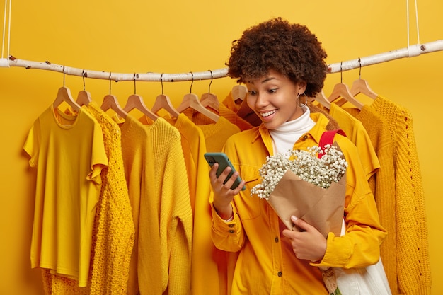 Foto de mujer feliz se viste para la primera cita, se para cerca del perchero de la ropa, recibe sms agradables en el teléfono inteligente, sostiene un hermoso ramo