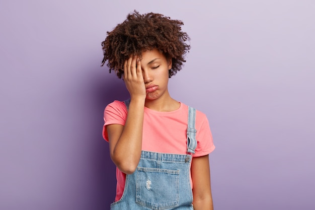 Foto de mujer frustrada y cansada de piel oscura que cubre la cara con la palma de la mano, se siente con exceso de trabajo, está preparada para el examen toda la noche, tiene expresión soñolienta, vestida con ropa elegante, modelos sobre una pared violeta