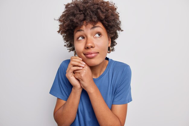 Foto de mujer guapa con cabello afro mantiene las manos cerca de la cara mira hacia arriba con expresión soñadora piensa en algo hace planes en mente weas camiseta casual aislada sobre pared blanca de estudio