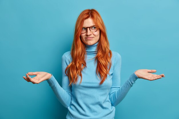 La foto de una mujer guapa indiferente con el pelo largo de color rojo se extiende por las palmas de las manos y parece desorientada, no puede decidir qué hacer, viste ropa informal.