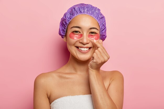 La foto de una mujer hermosa hace un signo de mano coreano, expresa amor, muestra el gesto del corazón con el dedo, usa gorro de baño, se para envuelto en una toalla, aplica parches cosméticos en los ojos, sonríe feliz.