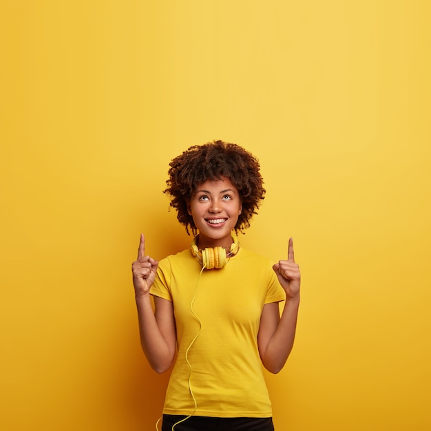 La foto de una mujer hipster sonriente apunta arriba con ambos dedos índices, muestra un lugar agradable en el piso de arriba, escucha la pista favorita en auriculares, usa una camiseta amarilla brillante en un tono con la pared