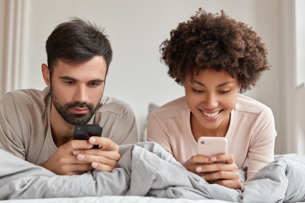 Foto de mujer y hombre adictos de diferentes razas, usar teléfonos móviles en la cama, conectarse a internet inalámbrico, disfrutar de la mañana los fines de semana, tener expresiones alegres. Concepto de tecnologías modernas