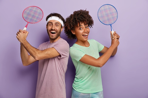 Foto de una mujer y un hombre alegres de raza mixta que están juntos, sostienen la raqueta de tenis, disfrutan jugando, ríen positivamente, se visten con camisetas, se mantienen activos y satisfechos