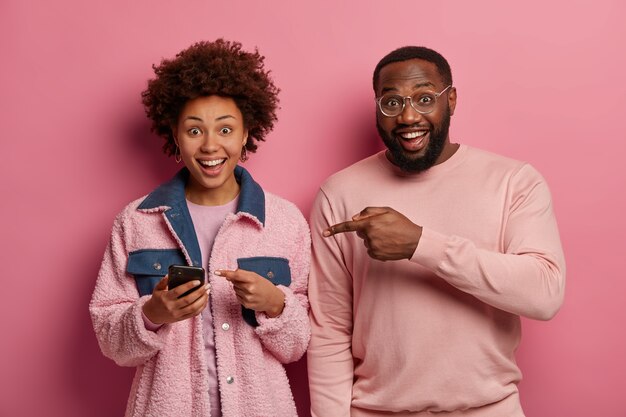 La foto de la mujer y el hombre alegres señalan el dispositivo del teléfono inteligente, miran contenido de video interesante, se colocan uno al lado del otro, sonríen ampliamente