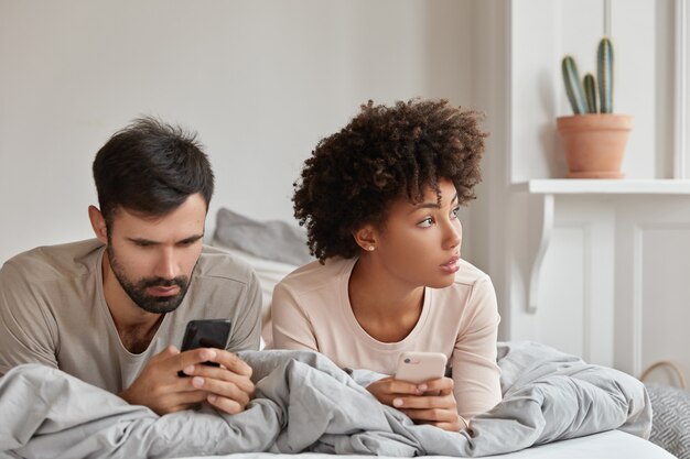 Foto de una mujer y un hombre de raza mixta adictos pensativos que leen el sitio web a través de la aplicación del teléfono móvil, pasan tiempo en la cama durante el sábado por la mañana, tienen adicción a las tecnologías. Par navegar por internet