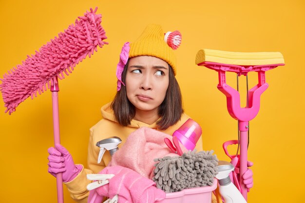 Foto de mujer insatisfecha concentrada con el ceño fruncido involucrado en el servicio de limpieza sostiene trapeadores para lavar el piso que va a lavar la ropa isoated sobre una pared amarilla