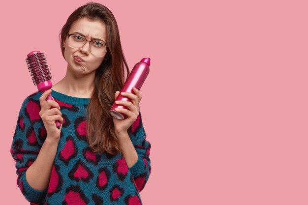 Foto de mujer insatisfecha frunce el ceño, se siente disgustada, lleva peine y laca para cortar el cabello, vestida con ropa doméstica