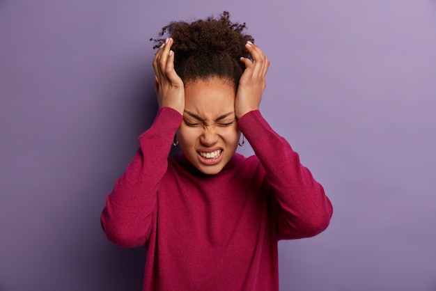 La foto de una mujer insatisfecha mantiene las manos en la sien, sufre de un dolor de cabeza insoportable, aprieta los dientes con dolor, está exhausta después del trabajo, usa un cuello alto de color burdeos, aislado en una pared púrpura.
