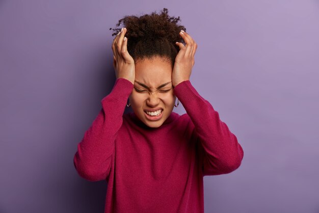 La foto de una mujer insatisfecha mantiene las manos en la sien, sufre de un dolor de cabeza insoportable, aprieta los dientes con dolor, está exhausta después del trabajo, usa un cuello alto de color burdeos, aislado en una pared púrpura.
