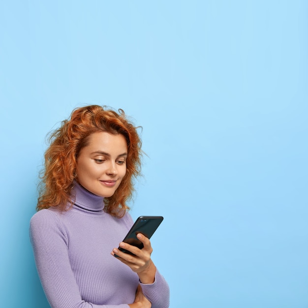 La foto de una mujer de jengibre de aspecto agradable está medio girada, usa un teléfono inteligente moderno, verifica la casilla de correo electrónico, vestida con ropa informal, aislada en la pared azul, copia espacio para publicidad