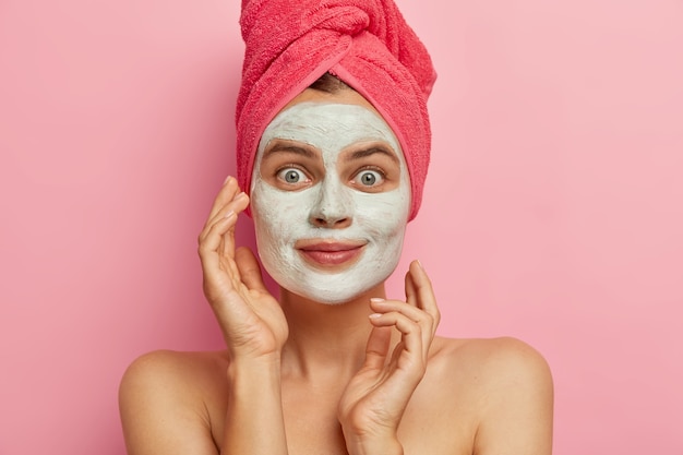 Foto de una mujer joven activa que aplica una mascarilla nutritiva para el cuidado facial, quiere tener la piel limpia y fresca, usa una toalla rosa en la cabeza