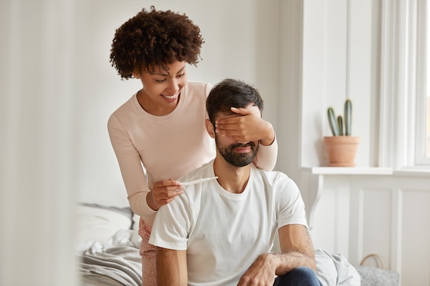 Foto de mujer joven alegre y alegre de piel oscura que cubre los ojos de su marido, quiere hacer una sorpresa inesperada, muestra un resultado positivo del embarazo, posa en la cama y se convierte en padre. Concepto de maternidad