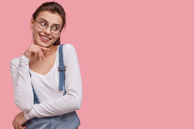 Foto de mujer joven atractiva con sonrisa agradable, inclina la cabeza, mantiene las manos parcialmente cruzadas, usa gafas, ropa de moda