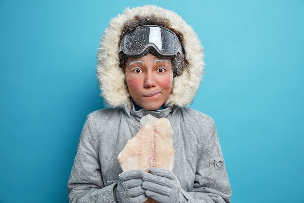 Foto de mujer joven congelada con cara roja usa chaqueta abrigada para las condiciones frías del invierno sostiene filete de pescado que necesita calor.