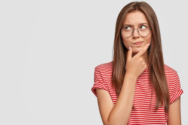 Foto de mujer joven dudosa insegura sostiene la barbilla, mira a la derecha con duda, se siente indeciso