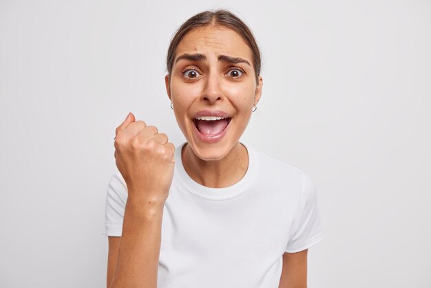 Foto de mujer joven emocional exlcaims frunce el ceño en voz alta cara aprieta puños reacciona ante noticias negativas promete venganza vestida informalmente aislada sobre pared blanca