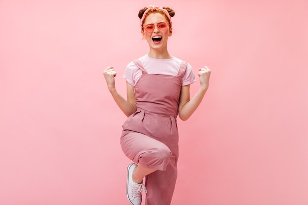 Foto de mujer joven de jengibre en gafas de sol en forma de corazones. Mujer en vestido y top blanco se ríe sobre fondo rosa.